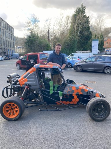 A person stands by a black and orange go-kart in a parking area with cars in the background.