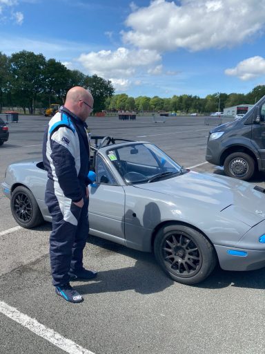 A man in racing gear stands beside a silver sports car under a blue sky.